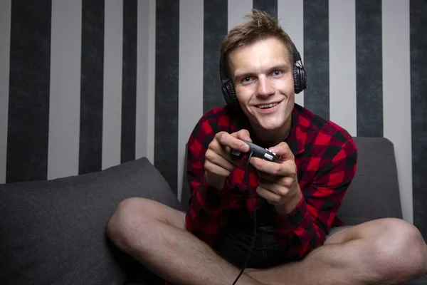 Jovem cara joga jogos de vídeo no console, ele se senta em casa no sofá e segura o joystick à noite — Fotografia de Stock