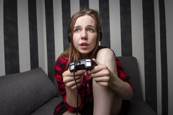 Jovem menina senta-se no sofá e joga o console no joystick, ela está focada em jogos de vídeo — Fotografia de Stock