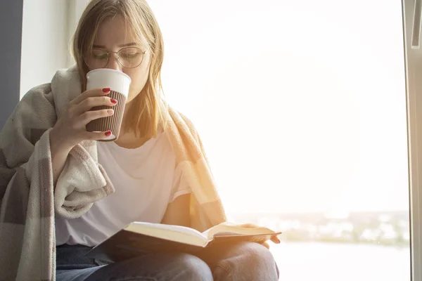 Jeune fille couverte d'une couverture se trouve à la maison par la fenêtre, lit un livre et boit du thé, espace de copie pour le texte — Photo