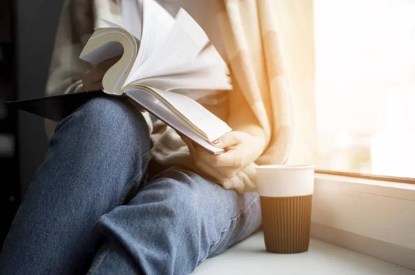 Jeune fille est assise près de la fenêtre, couverte d'une couverture, elle lit un livre et boit du thé, gros plan — Photo