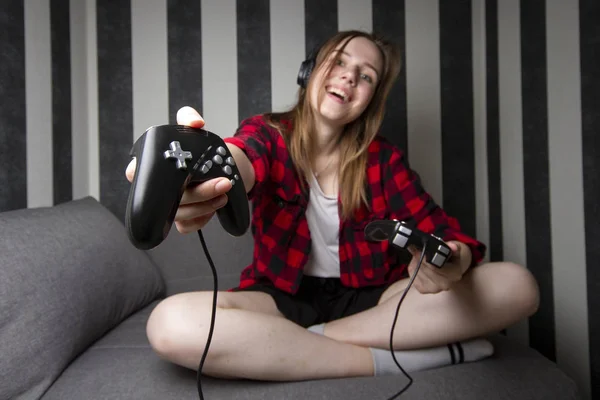 Uma jovem se senta sozinha em um sofá e joga videogames, ela segura um joystick e se oferece para jogar, ela está sozinha e mostra o dispositivo na câmera — Fotografia de Stock