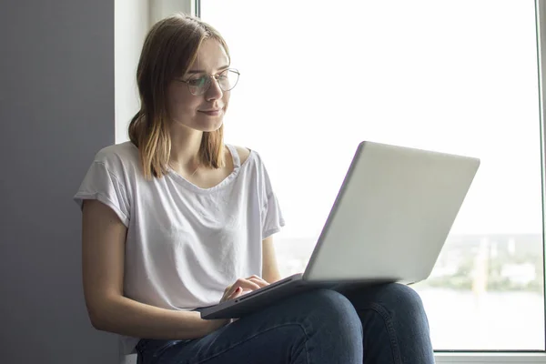 Jovem sentada perto da janela com um laptop, ela usa o computador em casa no peitoril da janela, uma mulher freelance, espaço de cópia para texto — Fotografia de Stock