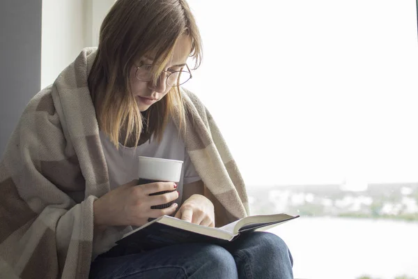 Jeune fille couverte d'une couverture se trouve à la maison par la fenêtre, lit un livre et boit du thé, espace de copie pour le texte — Photo