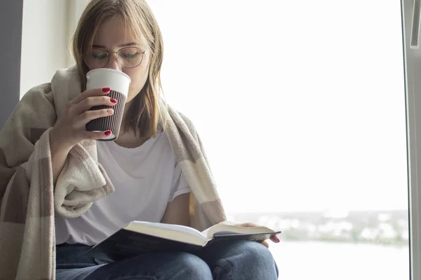 Jeune fille couverte d'une couverture se trouve à la maison par la fenêtre, lit un livre et boit du thé, espace de copie pour le texte — Photo