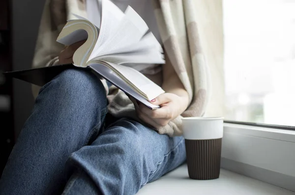 Молодая девушка, покрытая одеялом, сидит дома у окна, читает книгу и пьет чай, копирует место для текста — стоковое фото