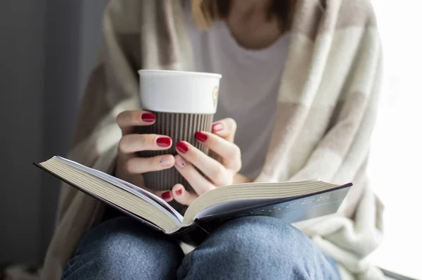 Jeune fille couverte d'une couverture se trouve à la maison par la fenêtre, lit un livre et boit du thé, espace de copie pour le texte — Photo