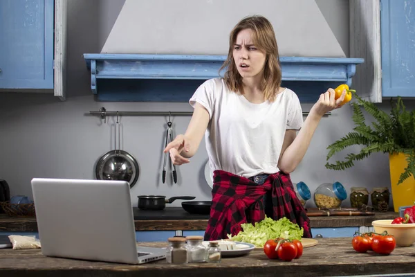 어린 소녀는 부엌에서 채식 샐러드를 준비하고 노트북을 들여다 보고, 건강한 음식을 준비하는 비디오 자습서로 요리하는 법을 배웁니다. — 스톡 사진
