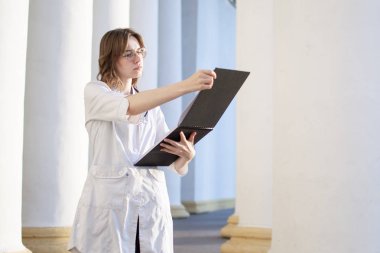 Genç bir hemşirenin portresi, bir medikal üniversite öğrencisi bir fonendoskop ile duruyor ve hastane kontrolleri belgelerinin arka planına karşı üniforma içinde mutlu kadın doktor okur