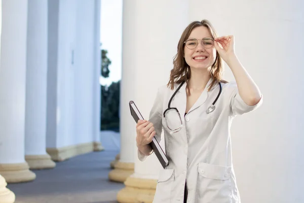 portrait of a young nurse, medical university student girl stands with phonendoscope and documents, happy female doctor in uniform near the hospital