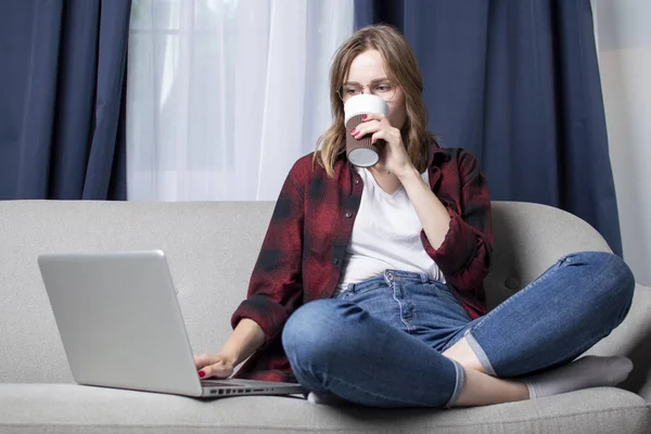 Girl is sitting on the couch with a laptop and is watching a video, female freelancer works at home, she uses a computer, copy space — Stock Photo, Image