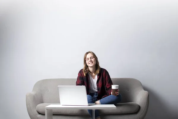 ソファに座ってコーヒーを飲むノートパソコンを持つ若い女の子、白い空白の壁に対してコンピュータを使用している女性、彼女はフリーランスと夢を見て、スペースをコピー — ストック写真
