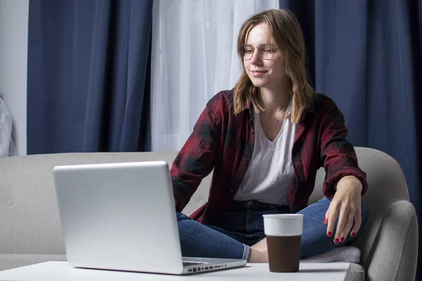 Girl is sitting on the couch with a laptop and is watching a video, female freelancer works at home, she uses a computer, copy space — Stock Photo, Image