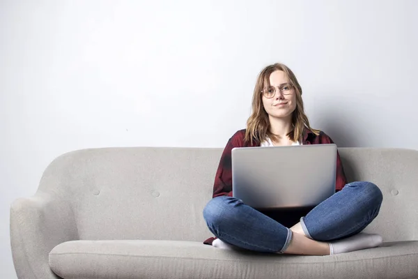 Jovem com um laptop sentado no sofá e beber café, uma mulher usando um computador contra uma parede branca em branco, ela freelancer e sonhando, espaço de cópia — Fotografia de Stock