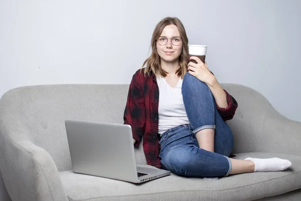 ソファに座ってコーヒーを飲むノートパソコンを持つ若い女の子、白い空白の壁に対してコンピュータを使用している女性、彼女はフリーランスとカメラを見て、スペースをコピー — ストック写真