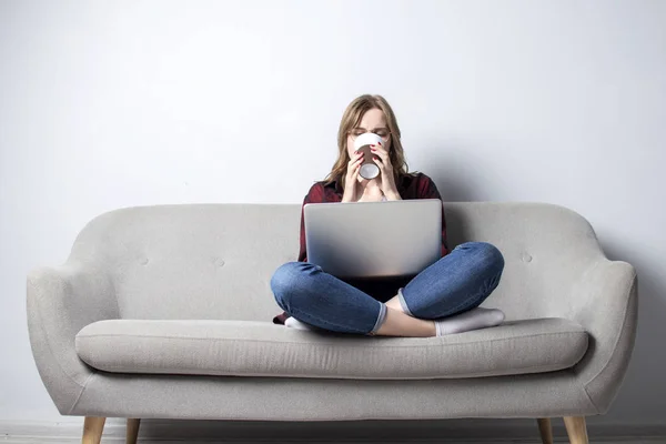 Gadis muda dengan laptop duduk di sofa dan minum kopi, seorang wanita menggunakan komputer terhadap dinding kosong putih, dia freelancing dan minum kopi, menyalin ruang — Stok Foto