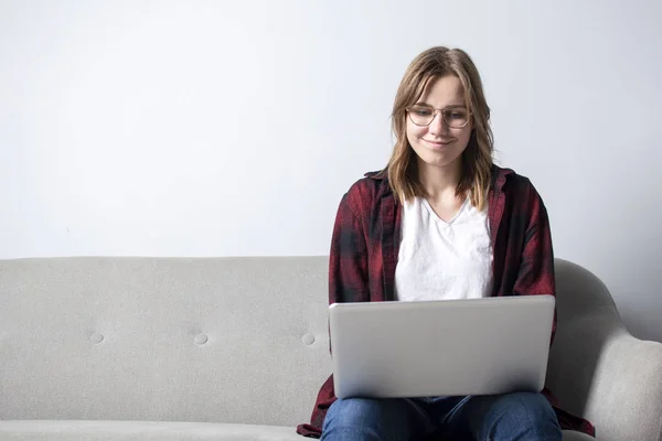 Jovem com um laptop sentado no sofá e sorrindo, uma mulher usando um computador contra uma parede branca em branco, ela freelancer e impressão de texto, espaço de cópia — Fotografia de Stock