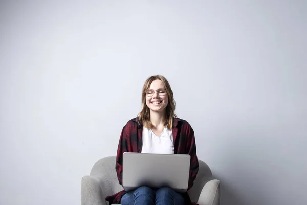 柔らかい快適な椅子に座ってラップトップを持つ若い女の子、およびスミリング、白い空白の壁に対してコンピュータを使用して女性、彼女はフリーランスとテキストを印刷し、スペースをコピー — ストック写真