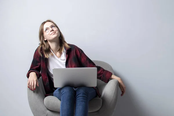 Jovem com um laptop sentado em uma cadeira macia confortável e pensando, uma mulher usando um computador contra uma parede branca em branco, ela freelancer e sonhando, espaço de cópia — Fotografia de Stock
