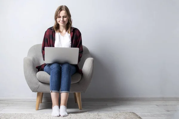 Jovem com um laptop sentado em uma cadeira macia confortável, e smilling, uma mulher usando um computador contra uma parede branca em branco, ela freelancer e texto de impressão, espaço de cópia — Fotografia de Stock