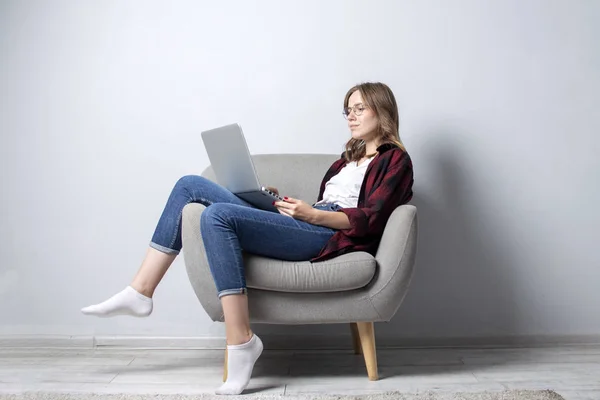Ung flicka med en bärbar dator sitter på en mjuk bekväm stol, och slappna av, en kvinna som använder en dator mot en vit blank vägg, frilansar hon och skriva ut text, kopiera utrymme — Stockfoto