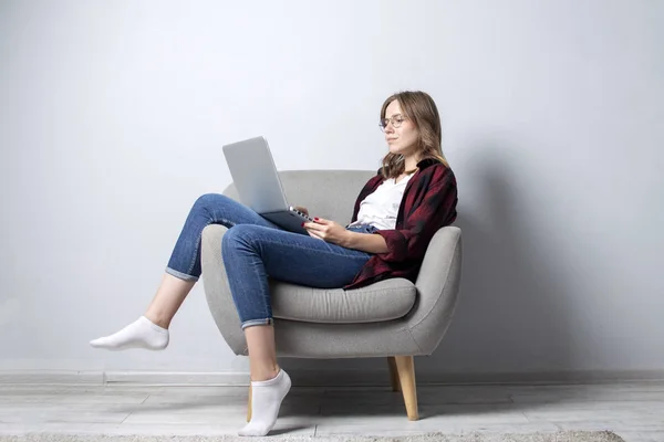 Muchacha joven con un ordenador portátil sentado en una silla cómoda suave, y relajarse, una mujer que usa una computadora contra una pared blanca en blanco, ella freelancing e imprimir texto, espacio de copia —  Fotos de Stock