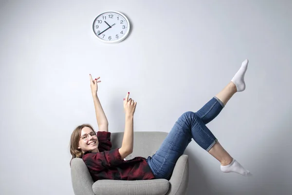 vacation is over. Young girl is sitting on a soft comfortable chair against a white wall and indicates the time, a hipster student is resting and dreaming, copy space