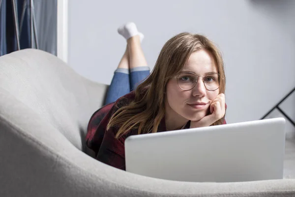 Pige ligger på sofaen med en bærbar computer og ser en video, kvindelig freelancer arbejder hjemme, hun bruger en computer - Stock-foto