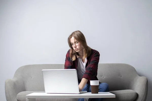 Jong meisje met een laptop zittend op de Bank en het drinken van koffie, een vrouw met behulp van een computer tegen een witte Blanco muur, ze Freelancing en afdrukken van tekst, kopieer ruimte — Stockfoto