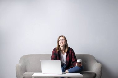 kanepede oturan ve kahve içen bir dizüstü bilgisayar ile genç kız, beyaz boş bir duvara karşı bir bilgisayar kullanan bir kadın, o freelancing ve rüya, kopya alanı