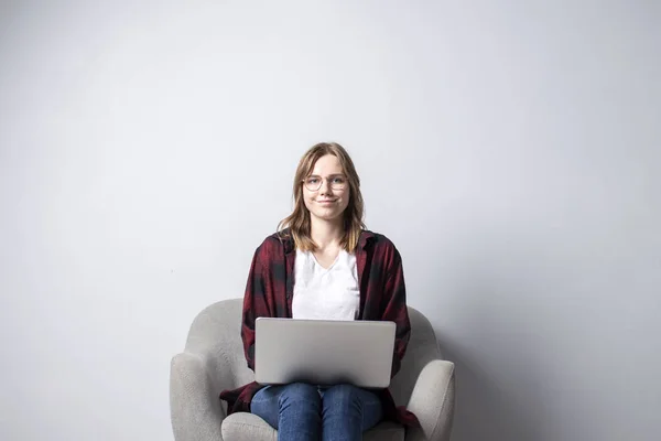 Jovem com um laptop sentado em uma cadeira macia confortável, e smilling, uma mulher usando um computador contra uma parede branca em branco, ela freelancer e texto de impressão, espaço de cópia — Fotografia de Stock