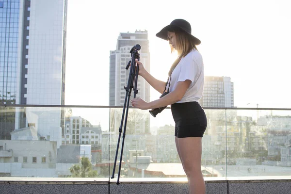 街を背景にカメラと三脚を持つ少女写真家、彼女は夕日を撮影し、撮影の準備をしている女性は、三脚を設定します — ストック写真