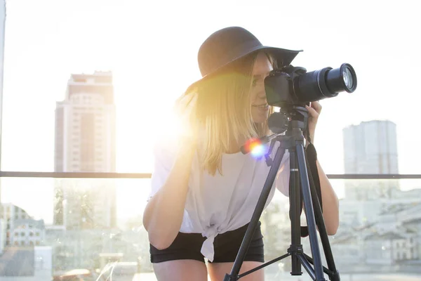 Fille photographe avec un appareil photo et un trépied sur un fond de ville, elle photographie au coucher du soleil, une femme tourne une vidéo — Photo