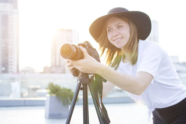 Fille photographe avec un appareil photo et un trépied sur un fond de ville, elle photographie au coucher du soleil, une femme tourne une vidéo — Photo