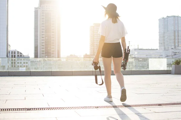 Joven fotógrafa con una cámara y un trípode sobre el fondo de la ciudad, fotografía la puesta de sol —  Fotos de Stock