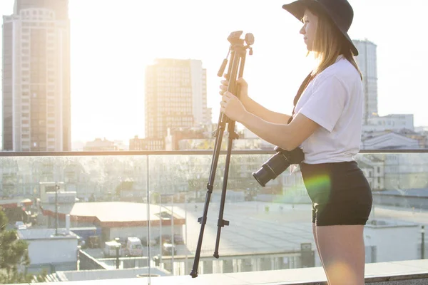 Chica-fotógrafa con una cámara y un trípode sobre el fondo de la ciudad, fotografía la puesta del sol, una mujer preparándose para disparar, monta un trípode —  Fotos de Stock