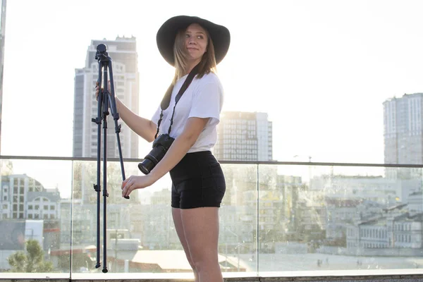 Tyttö-valokuvaaja, jolla on kamera ja kolmijalka kaupungin taustalla, hän kuvaa auringonlaskua, naista, joka valmistautuu ampumaan, perustaa kolmijalan. — kuvapankkivalokuva