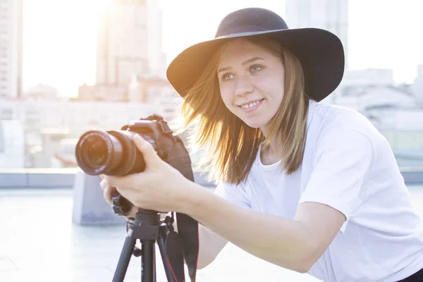 Fotógrafa con una cámara y un trípode sobre un fondo de la ciudad, fotografía al atardecer, una mujer graba un vídeo —  Fotos de Stock