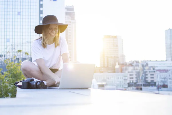 Fotógrafo menina sentado com câmera e laptop ao ar livre e retoque foto, freelancer turístico em um fundo de pôr do sol da cidade — Fotografia de Stock