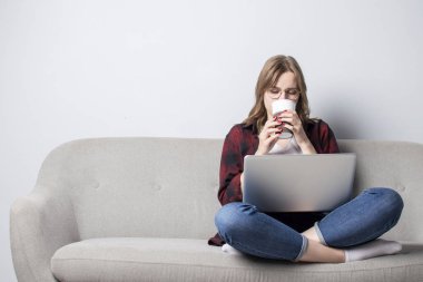 kanepede oturan ve kahve içen bir dizüstü bilgisayar ile genç kız, beyaz boş duvara karşı bir bilgisayar kullanan bir kadın, o freelancing ve kahve içme, kopya alanı