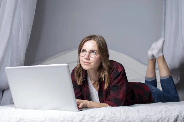 女の子はラップトップでベッドに横たわって、ビデオを見て、女性フリーランサーは自宅で働いて、彼女はコンピュータを使用し、コーヒーを飲んで、スペースをコピー — ストック写真