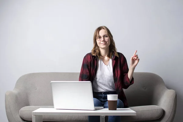 ソファに座ってコーヒーを飲んでいるノートパソコンを持つ若い女の子、白い空白の壁に対してコンピュータを使用している女性、彼女はフリーランスとコピースペースに指を指さす — ストック写真