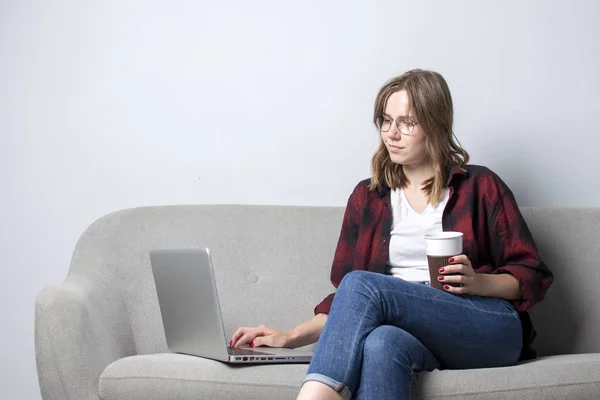 ソファに座ってコーヒーを飲むノートパソコンを持つ若い女の子、白い空白の壁に対してコンピュータを使用している女性、彼女はフリーランスとコーヒーを飲んで、スペースをコピー — ストック写真