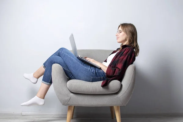 Jovem com um laptop sentado em uma cadeira macia confortável e smilling, uma mulher usando um computador contra uma parede branca em branco, ela freelancer e texto de impressão, espaço de cópia — Fotografia de Stock