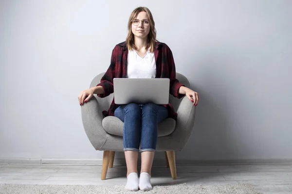Junges Mädchen mit Laptop, das auf einem weichen bequemen Stuhl sitzt und lächelt, eine Frau, die einen Computer vor einer weißen leeren Wand benutzt, sie arbeitet freiberuflich und schaut in die Kamera, Kopierraum — Stockfoto