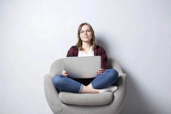 Jovem com um laptop sentado em uma cadeira macia confortável, e smilling, uma mulher usando um computador contra uma parede branca em branco, ela freelancer e texto de impressão, espaço de cópia — Fotografia de Stock