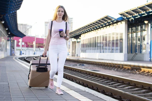 attractive girl is standing with luggage at the station and waiting for the train, the student is going on a trip, she is walking along the platform, copy space