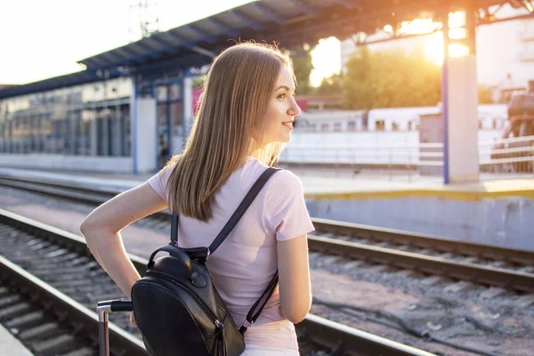 Chica se para en la plataforma de la estación con una maleta y espera un tren, un estudiante viaja, sale de vacaciones de verano, espacio para copiar —  Fotos de Stock