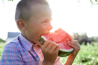 çocuk köyde yeşil çim oturur ve karpuz büyük bir parça tutar, küçük bir okul çocuğu güneşli bir yaz gününde bir karpuz yiyor, yakın çekim