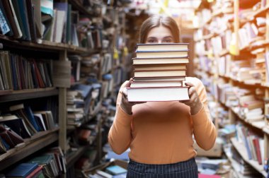 kız öğrenci kütüphanede kitap yığını tutar, o edebiyat arar ve okumak için teklif, bir kadın çalışma için hazırlar, bilgi güç, kitapçı arka planda kitapçı, kibir