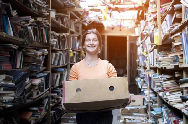 Девушка несет коробку с книгами в библиотеке, портрет продавца на фоне книжного магазина — стоковое фото